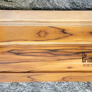 May include: A rectangular wooden serving tray with a light brown finish and dark brown accents. The tray has black handles on each end and the text "The Grillfather JOE'S BBQ" is engraved on the surface.
