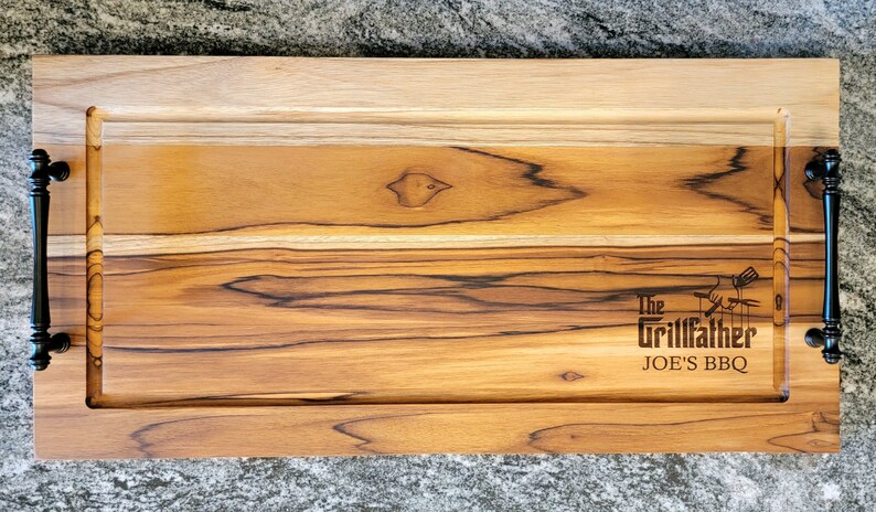 May include: A rectangular wooden serving tray with a light brown finish and dark brown accents. The tray has black handles on each end and the text "The Grillfather JOE'S BBQ" is engraved on the surface.