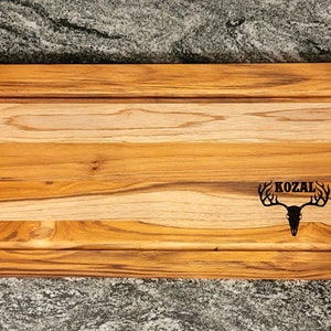 May include: A rectangular wooden serving board with a light brown finish, featuring a carved groove and black metal handles. The board is engraved with the word "KOZAL" above a deer skull design.