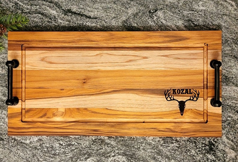 May include: A rectangular wooden serving board with a light brown finish, featuring a carved groove and black metal handles. The board is engraved with the word "KOZAL" above a deer skull design.