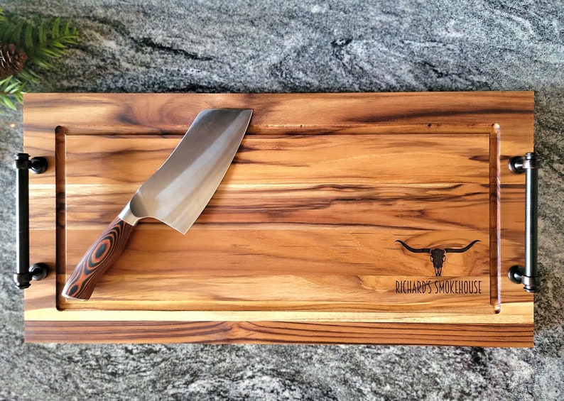 May include: A large wooden cutting board with a black metal handle on each end. The board has a groove around the perimeter and a smaller groove in the center. A silver cleaver with a black and red handle is resting on the board. The board is engraved with a bull skull and the text 'Richard's Smokehouse'.