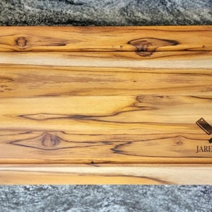 May include: A rectangular wooden cutting board with a light brown finish and dark brown handles. The board has a carved-out section and features the text "Jared's BBQ" with crossed knives.