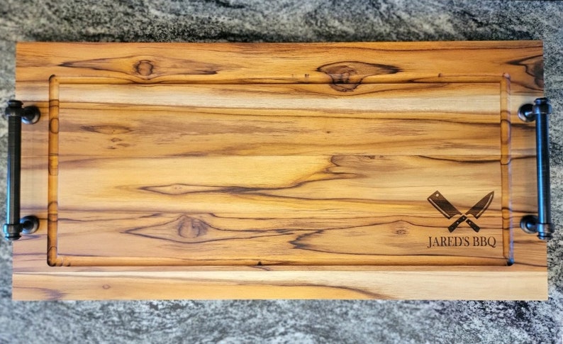 May include: A rectangular wooden cutting board with a light brown finish and dark brown handles. The board has a carved-out section and features the text "Jared's BBQ" with crossed knives.