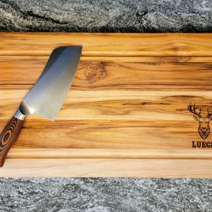 May include: A rectangular wooden cutting board with a stainless steel knife. The board has a carved-out section and black handles. The board is engraved with a deer head and the name "LUECK".