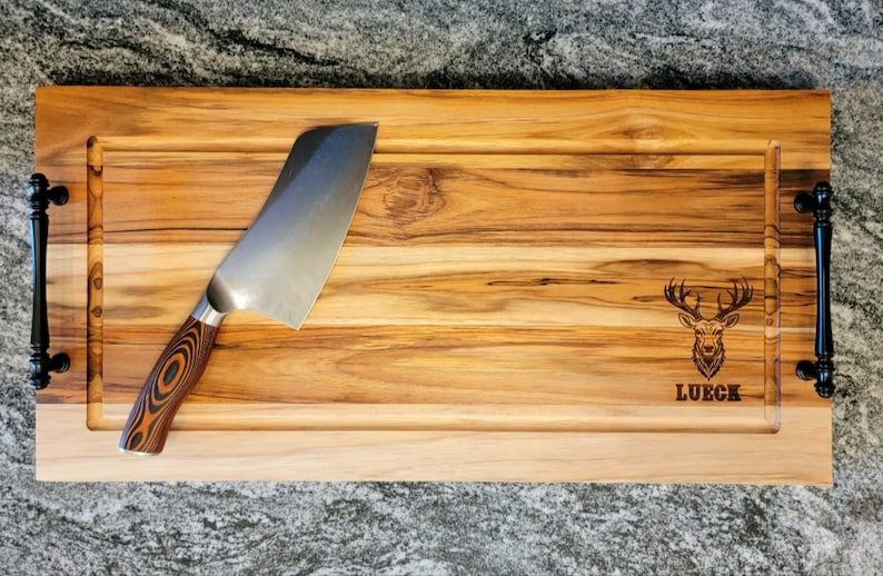 May include: A rectangular wooden cutting board with a stainless steel knife. The board has a carved-out section and black handles. The board is engraved with a deer head and the name "LUECK".