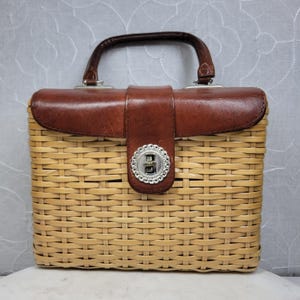 May include: A vintage rectangular handbag with a woven wicker body and a brown leather top and handle. The bag features a silver-toned clasp and a leather strap closure. The handle is curved and attached with silver hardware.