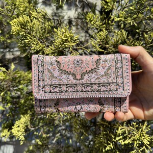 May include: A pink wallet with a patterned design that resembles a rug. The pattern is in shades of pink, white, and black.
