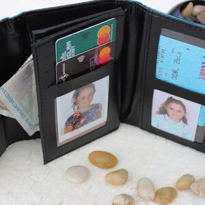 May include: A black leather wallet with multiple compartments, including slots for cards and cash, and two clear plastic windows for photos. The wallet is open and shows a few cards, cash, and two photos.