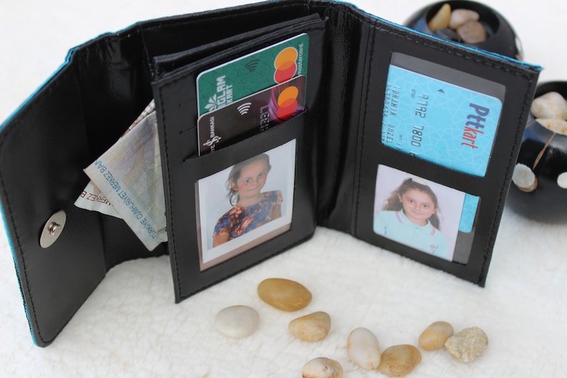 May include: A black leather wallet with multiple compartments, including slots for cards and cash, and two clear plastic windows for photos. The wallet is open and shows a few cards, cash, and two photos.