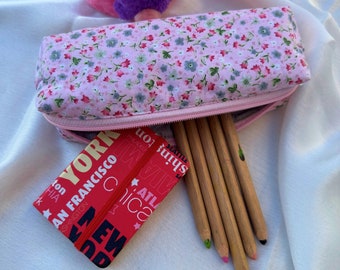 May include: A pink floral pencil case with a zipper, partially open to reveal several pencils. A small red notebook with white text, including city names like "New York" and "San Francisco", is next to the case. The pencils are wooden with colored tips.