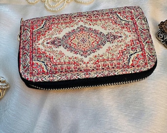 May include: A rectangular wallet featuring a detailed woven pattern in shades of red, cream, and black, reminiscent of a traditional rug design. The wallet is secured with a black zipper that runs along the edges, providing a contrast to the intricate textile surface.