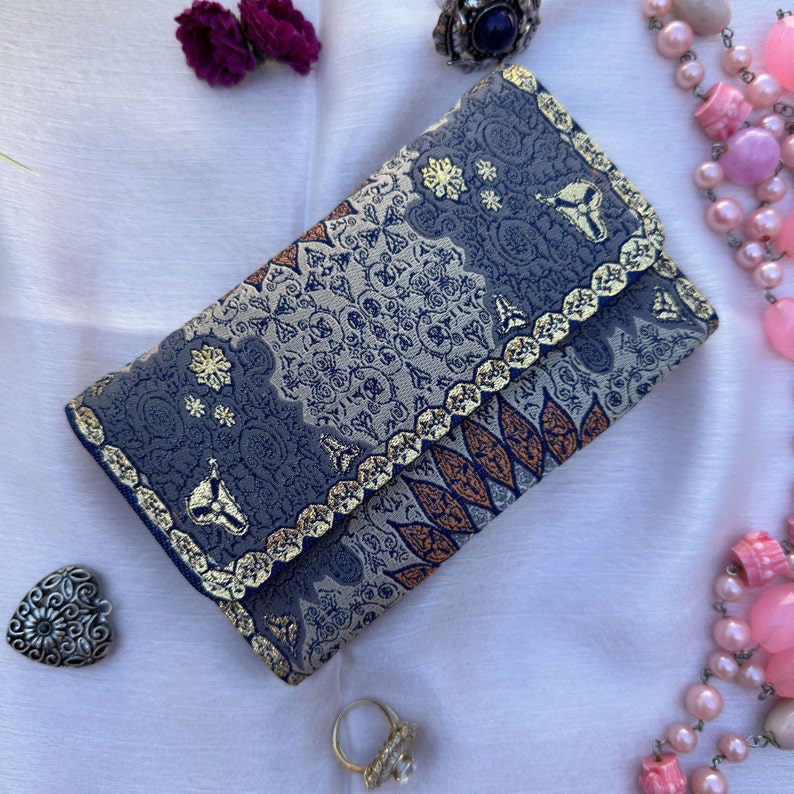 May include: A blue and gold patterned clutch purse with a gold trim. The purse has a floral design and is made of a soft fabric.