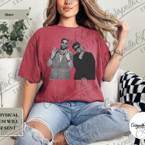 May include: A maroon-colored shirt featuring a black and white image of two men. The shirt has the word "Shirt" in a banner at the top left. The text "PHYSICAL ITEM WILL BE SENT" is at the bottom left.