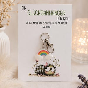 Könnte beinhalten: Eine weiße Karte mit deutschem Text und einem Schlüsselanhänger. Der Schlüsselanhänger zeigt einen Regenbogen und eine Cartoon-Raupe. Auf der Karte steht "EIN GLÜCKSANHÄNGER FÜR DICH" und "ER IST IMMER AN DEINER SEITE, WENN DU ES BRAUCHST!".