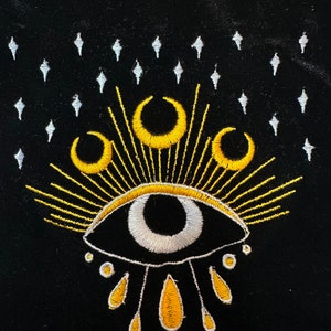 Puede incluir: Una bolsa de terciopelo negro con un ojo bordado en oro y blanco con tres lunas crecientes por encima y un sol radiante. El ojo tiene una pupila blanca y un iris negro. Hay cinco formas de l&aacute;grima amarillas debajo del ojo.