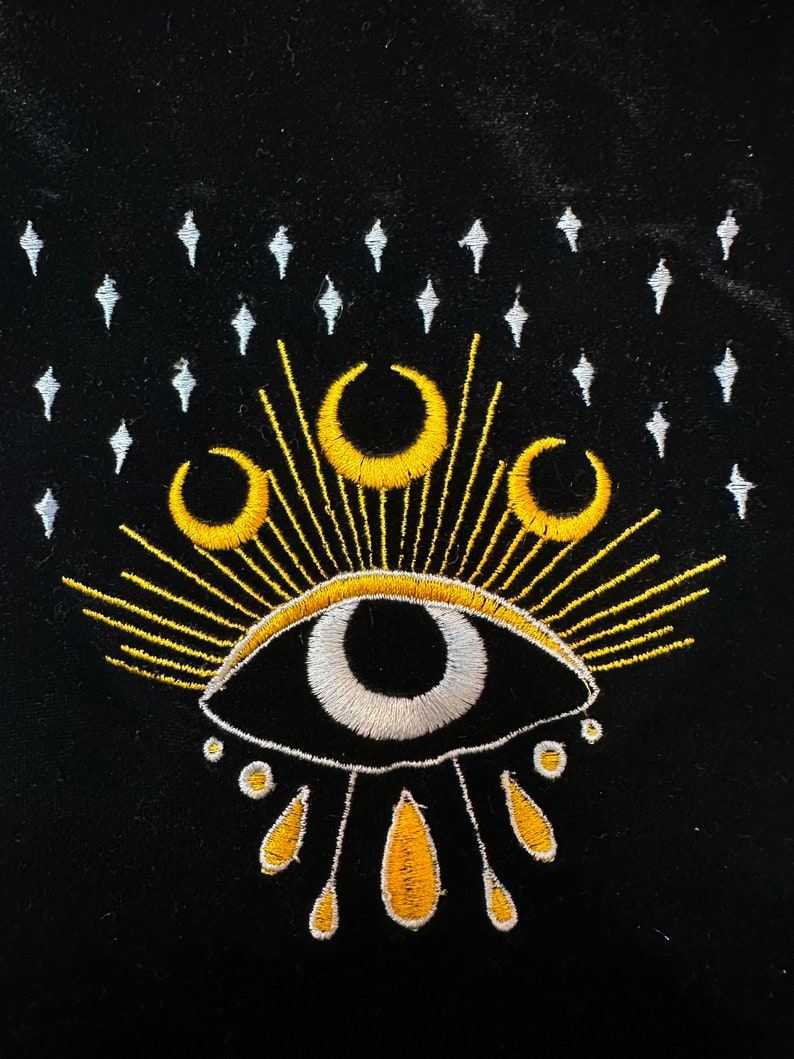 Puede incluir: Una bolsa de terciopelo negro con un ojo bordado en oro y blanco con tres lunas crecientes por encima y un sol radiante. El ojo tiene una pupila blanca y un iris negro. Hay cinco formas de l&aacute;grima amarillas debajo del ojo.