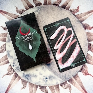 Könnte beinhalten: Ein schwarzes Orakelkarten-Deck und eine Karte mit dem Text "Wanderer's Oracle" mit einem grünen Blatt- und roten Halbmond-Design. Die Karte zeigt ein rosa Schlangenmuster auf schwarzem Hintergrund. Das Deck und die Karte befinden sich auf einer kreisförmigen Oberfläche.