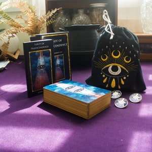 Puede incluir: Dos barajas de tarot, "Tarot of Transcendence" y "Tarot of Cendence", con una baraja de cartas azul y blanca delante de ellas. Una bolsa de cord&oacute;n negro con un ojo bordado en oro y lunas crecientes se encuentra junto a las cartas. Cuatro alfileres dorados con un dise&ntilde;o de estrella est&aacute;n sobre la mesa.