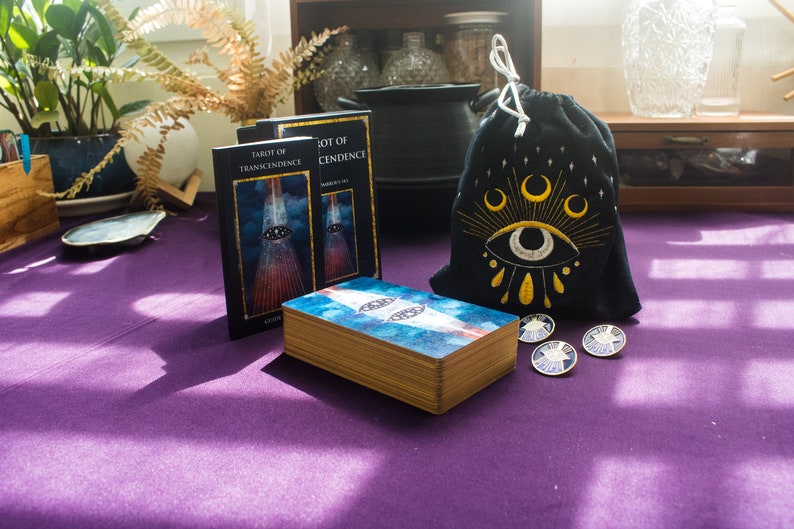 Puede incluir: Dos barajas de tarot, "Tarot of Transcendence" y "Tarot of Cendence", con una baraja de cartas azul y blanca delante de ellas. Una bolsa de cord&oacute;n negro con un ojo bordado en oro y lunas crecientes se encuentra junto a las cartas. Cuatro alfileres dorados con un dise&ntilde;o de estrella est&aacute;n sobre la mesa.