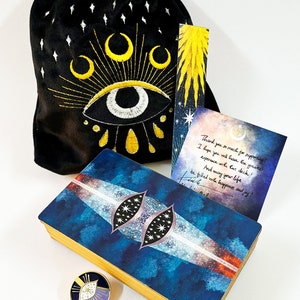 Puede incluir: Una baraja de tarot azul y dorada con un dise&ntilde;o de ojo celestial en la parte delantera. La baraja viene con una bolsa de terciopelo negro con un dise&ntilde;o de ojo bordado, un marcap&aacute;ginas con un dise&ntilde;o de estallido de estrellas amarillo y un peque&ntilde;o pin de esmalte con un dise&ntilde;o de ojo similar. La caja de la baraja tambi&eacute;n incluye una nota manuscrita que dice "&iexcl;Muchas gracias por tu apoyo! &iexcl;Espero que tengas la mejor experiencia con esta baraja! &iexcl;Y que tu vida est&eacute; llena de felicidad y alegr&iacute;a!"