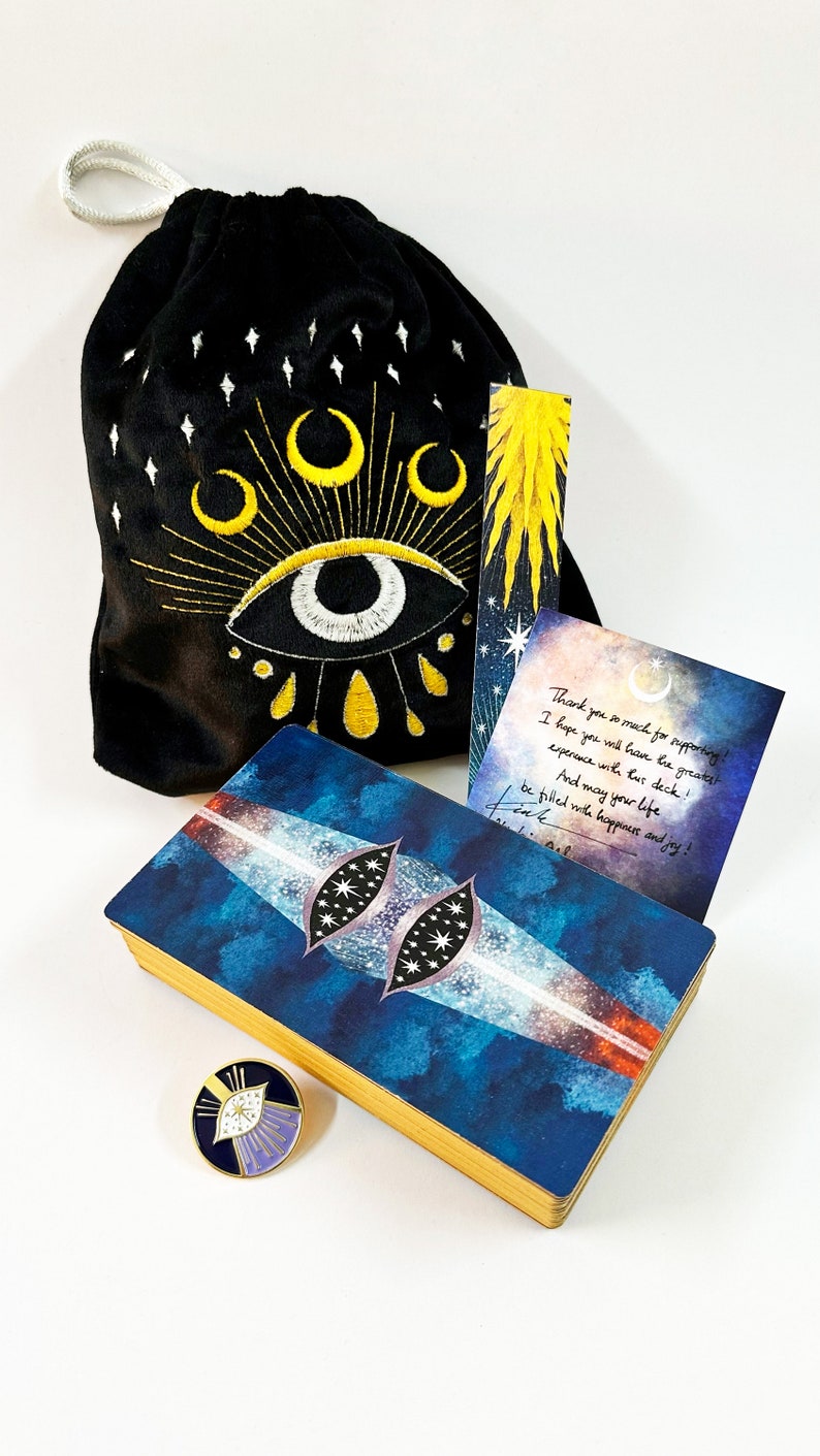 Puede incluir: Una baraja de tarot azul y dorada con un dise&ntilde;o de ojo celestial en la parte delantera. La baraja viene con una bolsa de terciopelo negro con un dise&ntilde;o de ojo bordado, un marcap&aacute;ginas con un dise&ntilde;o de estallido de estrellas amarillo y un peque&ntilde;o pin de esmalte con un dise&ntilde;o de ojo similar. La caja de la baraja tambi&eacute;n incluye una nota manuscrita que dice "&iexcl;Muchas gracias por tu apoyo! &iexcl;Espero que tengas la mejor experiencia con esta baraja! &iexcl;Y que tu vida est&eacute; llena de felicidad y alegr&iacute;a!"