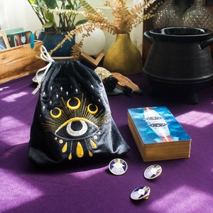 Puede incluir: Una bolsa de terciopelo negro con un ojo y lunas crecientes bordadas en oro. La bolsa est&aacute; sobre una superficie morada con tres alfileres dorados con dise&ntilde;os de ojos azules y blancos.