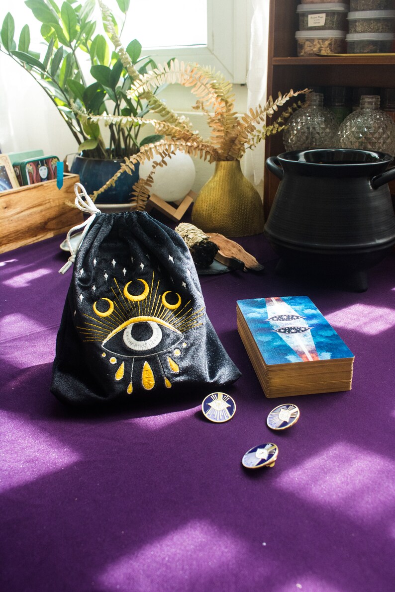 Puede incluir: Una bolsa de terciopelo negro con un ojo y lunas crecientes bordadas en oro. La bolsa est&aacute; sobre una superficie morada con tres alfileres dorados con dise&ntilde;os de ojos azules y blancos.