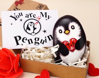 May include: A hand-painted penguin rock holding a red heart with the letters T+A, nestled in a brown box with white paper filler. A card reads You are My Penguin with a penguin graphic. A red fabric rose and felt hearts are nearby.