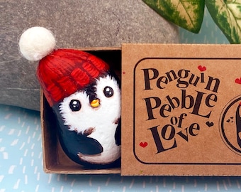 May include: A small painted penguin pebble wearing a red knit hat with a white pom-pom, nestled inside a brown cardboard matchbox. The box lid features the printed text Penguin Pebble of Love with small red heart icons and a circular graphic.