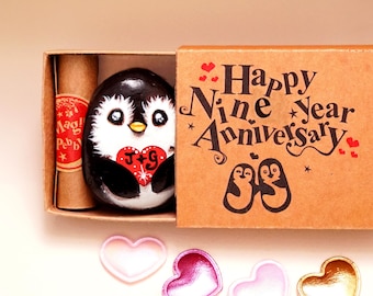 May include: A brown cardboard matchbox containing a painted black and white penguin rock holding a red heart with the letters J+G. The box lid reads Happy Nine Year Anniversary with two penguin illustrations. A scroll and four small hearts are included.