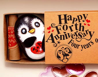 May include: A brown cardboard matchbox containing a painted penguin rock with a red heart labeled J+A and a small scroll. The box lid features the text Happy Forth Anniversary Four years of and a circular illustration of two penguins.