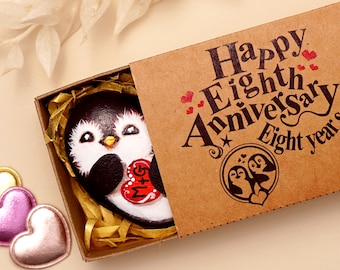 May include: A brown cardboard gift box containing a hand-painted black and white penguin rock holding a red heart with the letters M+G. The box lid features the text Happy Eighth Anniversary Eight years and a circular penguin illustration.