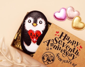 May include: A hand-painted black and white penguin rock holding a red heart with the letters A+K, nestled in a cardboard box. The box lid features the text Happy Seventh Anniversary Seven years of us! and a penguin illustration.