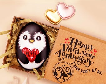 May include: A painted penguin rock holding a red heart with the letters H and R, nestled in a box with gold paper shreds. The box lid features the text Happy Third Year Anniversary Three years of us and two heart-shaped charms.