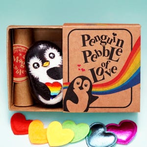 Puede incluir: Un guijarro de pingüino pintado en blanco y negro con un corazón arcoíris, dentro de una caja de cartón que dice "Penguin Pebble of Love". La caja también contiene un pergamino con la etiqueta "Magic Pebbles". Decoraciones coloridas en forma de corazón están dispuestas debajo.