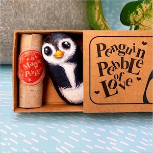 May include: A painted penguin pebble in a matchbox. The penguin is black, white, and orange. The box is printed with "Penguin Pebble of Love". A small scroll with "Magic Pebbles" is also included in the box.