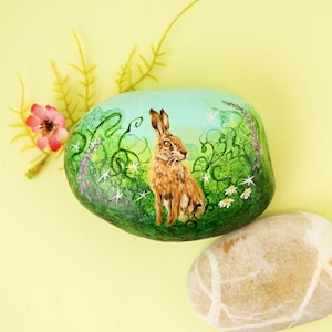 May include: A hand-painted, oval stone features a brown hare in a green meadow with white daisies and purple flowers. The background is a light blue sky. Additional decorative elements include a pink flower and yellow sprigs.