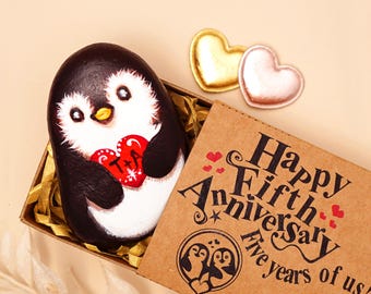 May include: A hand-painted penguin rock featuring a red heart with the letters T and A, nestled in a brown gift box. The box lid displays the text Happy Fifth Anniversary Five years of us and a penguin illustration with two small hearts.