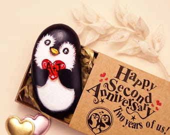 May include: A hand-painted black and white penguin rock holding a red heart with the letters A+J, nestled in a brown box. The box lid features the text Happy Second Anniversary Two years of us! and a graphic of two penguins with hearts.