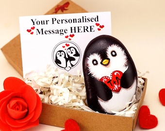 May include: A hand-painted penguin rock featuring a red heart with the letters T+A, nestled in a brown gift box with white paper filler. A card displays the text Your Personalised Message HERE and a circular penguin illustration with red hearts.