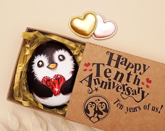 May include: A hand-painted penguin rock inside a brown gift box with gold filler. The penguin holds a red heart with the letters G and T. The box lid features the text Happy Tenth Anniversary Ten years of us and a penguin illustration.