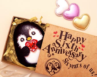 May include: A painted rock featuring a black and white penguin holding a red heart with the letters L and P, nestled in a box with the text Happy Sixth Anniversary Six years of us! and three small heart-shaped patches in pink, gold, and white.