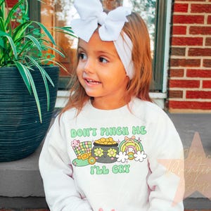 May include: A white long-sleeved shirt with a green, yellow, and gold graphic of a pot of gold and a leprechaun hat. The text "Don't Pinch Me I'll Cry" is printed on the shirt.