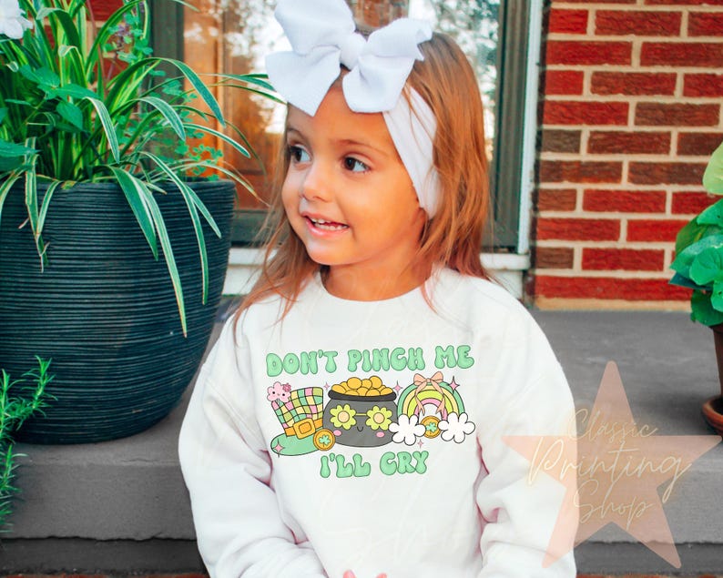 May include: A white long-sleeved shirt with a green, yellow, and gold graphic of a pot of gold and a leprechaun hat. The text "Don't Pinch Me I'll Cry" is printed on the shirt.
