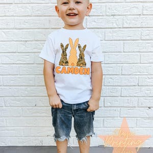 May include: A white t-shirt with a graphic of three brown and orange bunnies with the name "CAMDEN" printed below. A young boy is wearing the shirt and blue denim shorts.