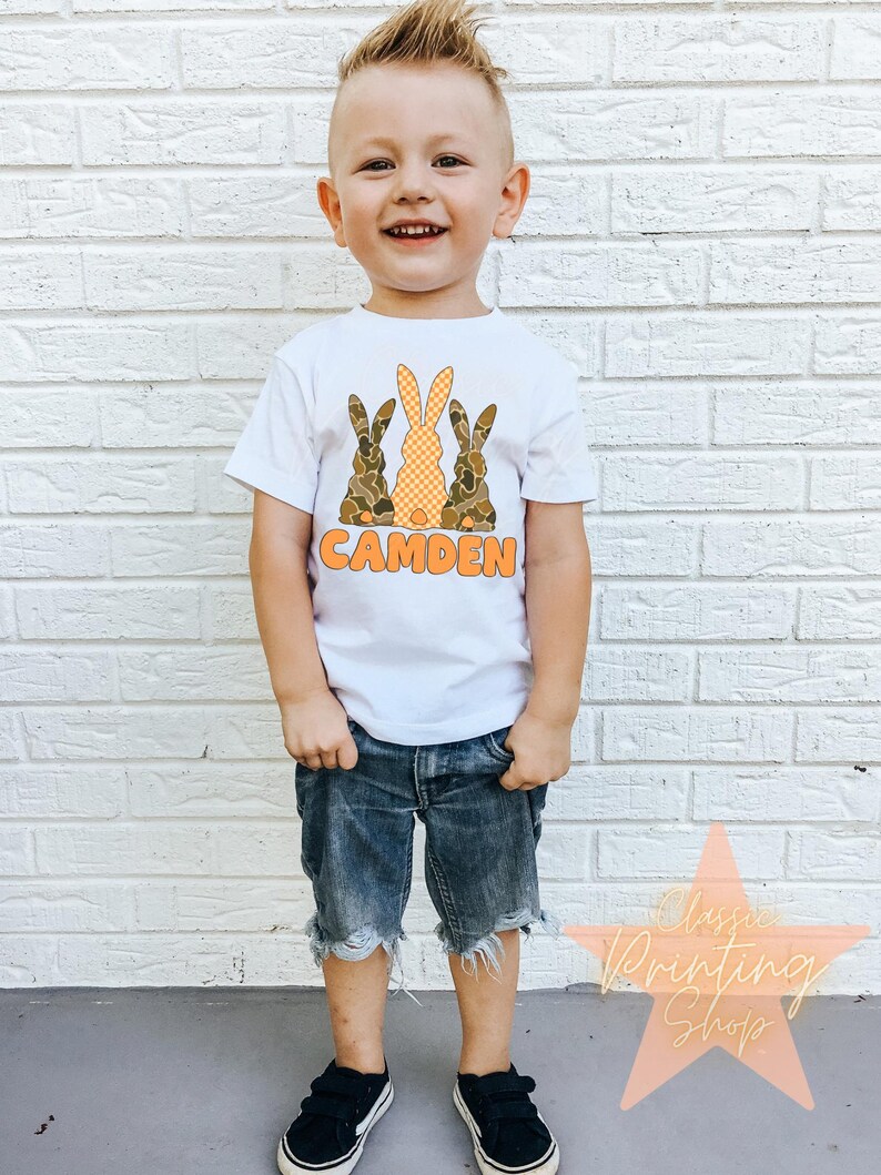 May include: A white t-shirt with a graphic of three brown and orange bunnies with the name "CAMDEN" printed below. A young boy is wearing the shirt and blue denim shorts.