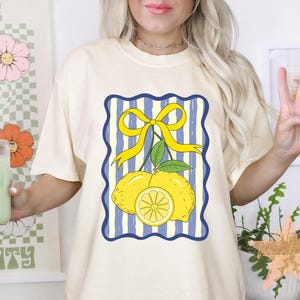 May include: Cream-colored t-shirt featuring a lemon graphic with a yellow bow and blue striped background. The graphic includes two whole lemons and a lemon slice. The shirt is a casual, everyday wear item.