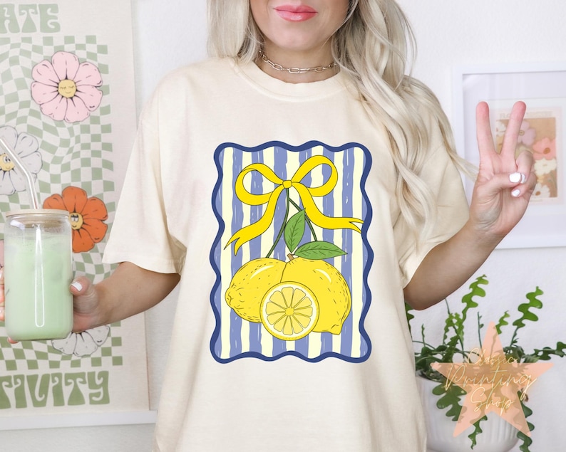 May include: Cream-colored t-shirt featuring a lemon graphic with a yellow bow and blue striped background. The graphic includes two whole lemons and a lemon slice. The shirt is a casual, everyday wear item.