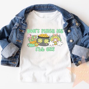 May include: White t-shirt with a green, yellow, and pink cartoon design. The text on the shirt reads "Don't Pinch Me I'll Cry".