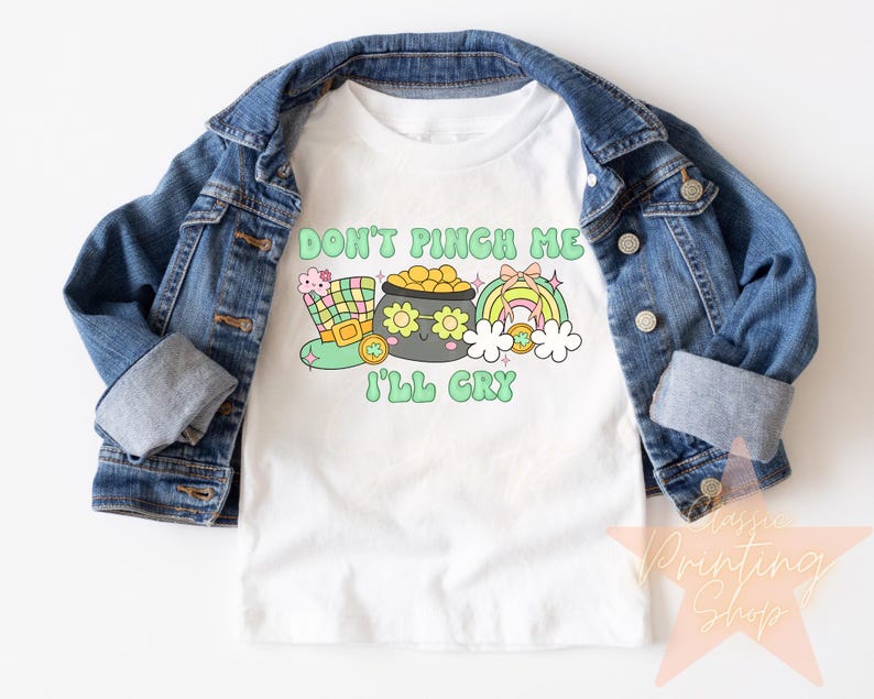 May include: White t-shirt with a green, yellow, and pink cartoon design. The text on the shirt reads "Don't Pinch Me I'll Cry".